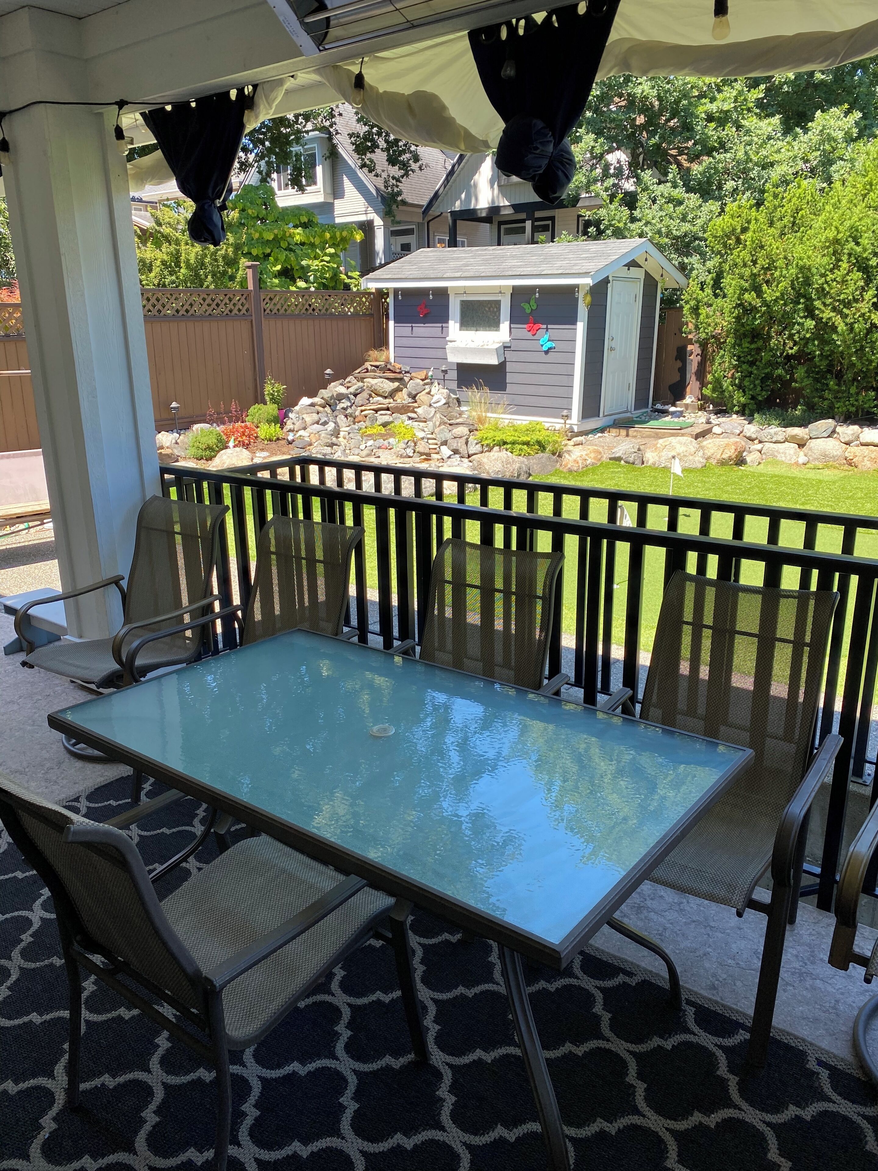 A covered patio area with a glass-top table and chairs, a small shed, and a garden with rocks and plants.