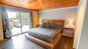 A bedroom with a wooden ceiling, a bed with grey bedding, two bedside lamps, and a sliding glass door leading to an outdoor patio with furniture. - Beautiful golf course getaway on Madeline island<br> (La Pointe)