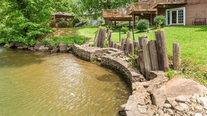 Un mur de soutènement en pierre, une clôture en bois, un bassin et une maison dotée d’une terrasse et d’un espace couvert.