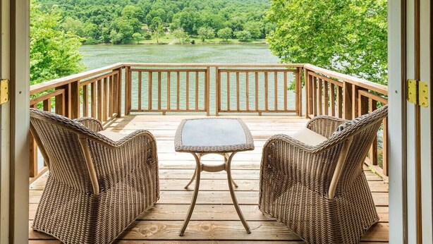 Une terrasse en bois, agrémentée de meubles en osier et d’une table en verre, offrant une vue sur une rivière et des collines boisées.