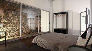 Een slaapkamer met een grote schuifglazen deur die uitkomt op een stenen muur, een bed met een beige sprei, een nachtkastje en een kapstok.