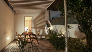 Een terras buiten met een houten tafel en stoelen, een lantaarn, en een trap naar een hoger gelegen niveau.