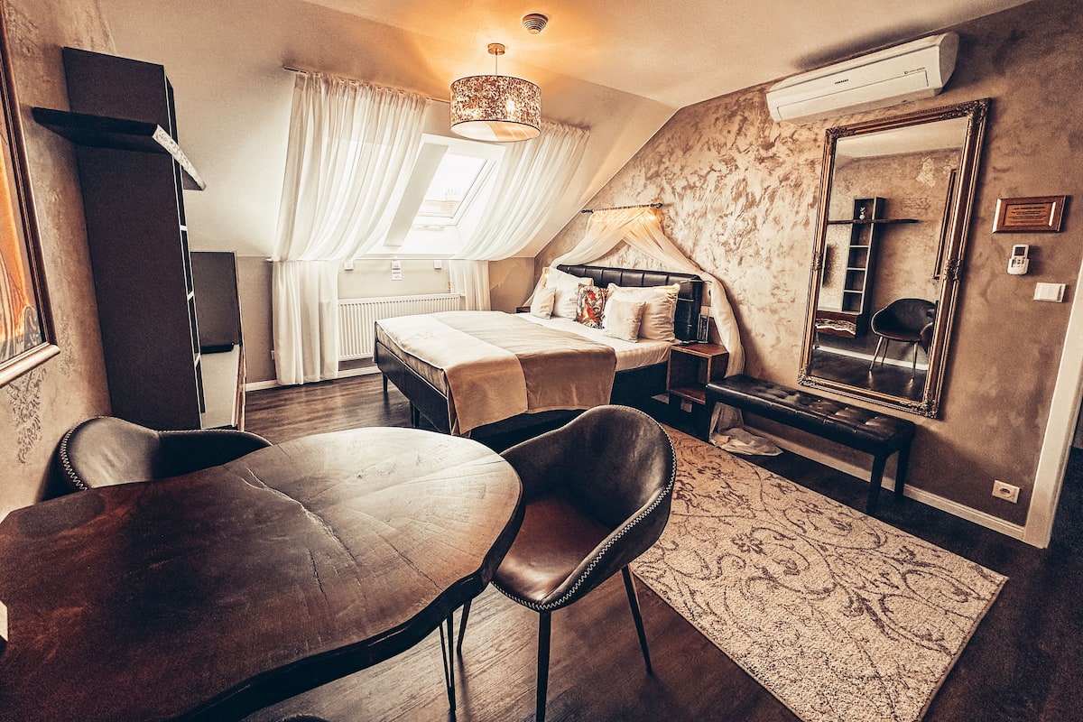 A bedroom with a bed, bedside table, lamp, mirror, and a view of the outdoors through a window.
