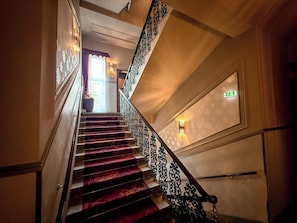 A grand staircase with ornate black railings and a red carpeted floor, leading to a landing with a door marked with an emergency exit sign.