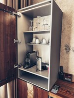 A cabinet with shelves containing glassware, cups, a coffee maker, and a menu sign.