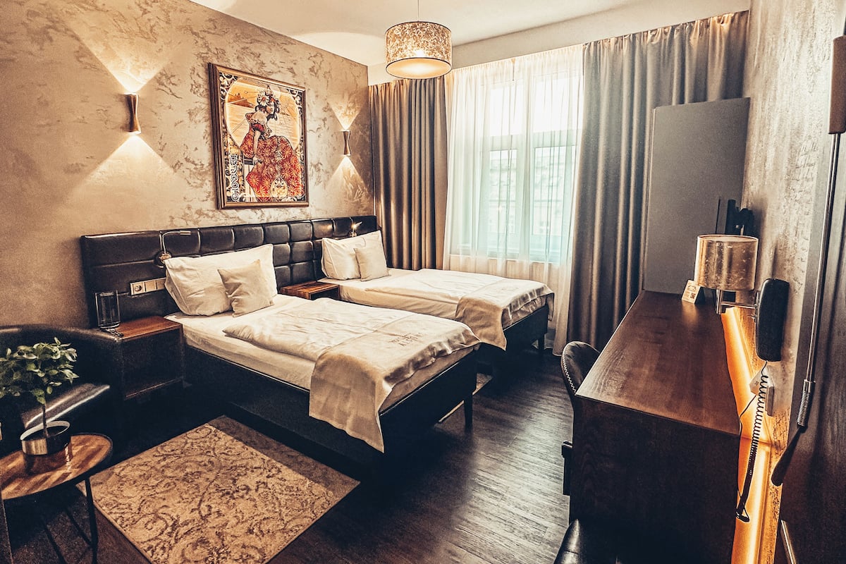 A hotel room with a double bed, a desk, a chair, a TV, and a window with curtains.