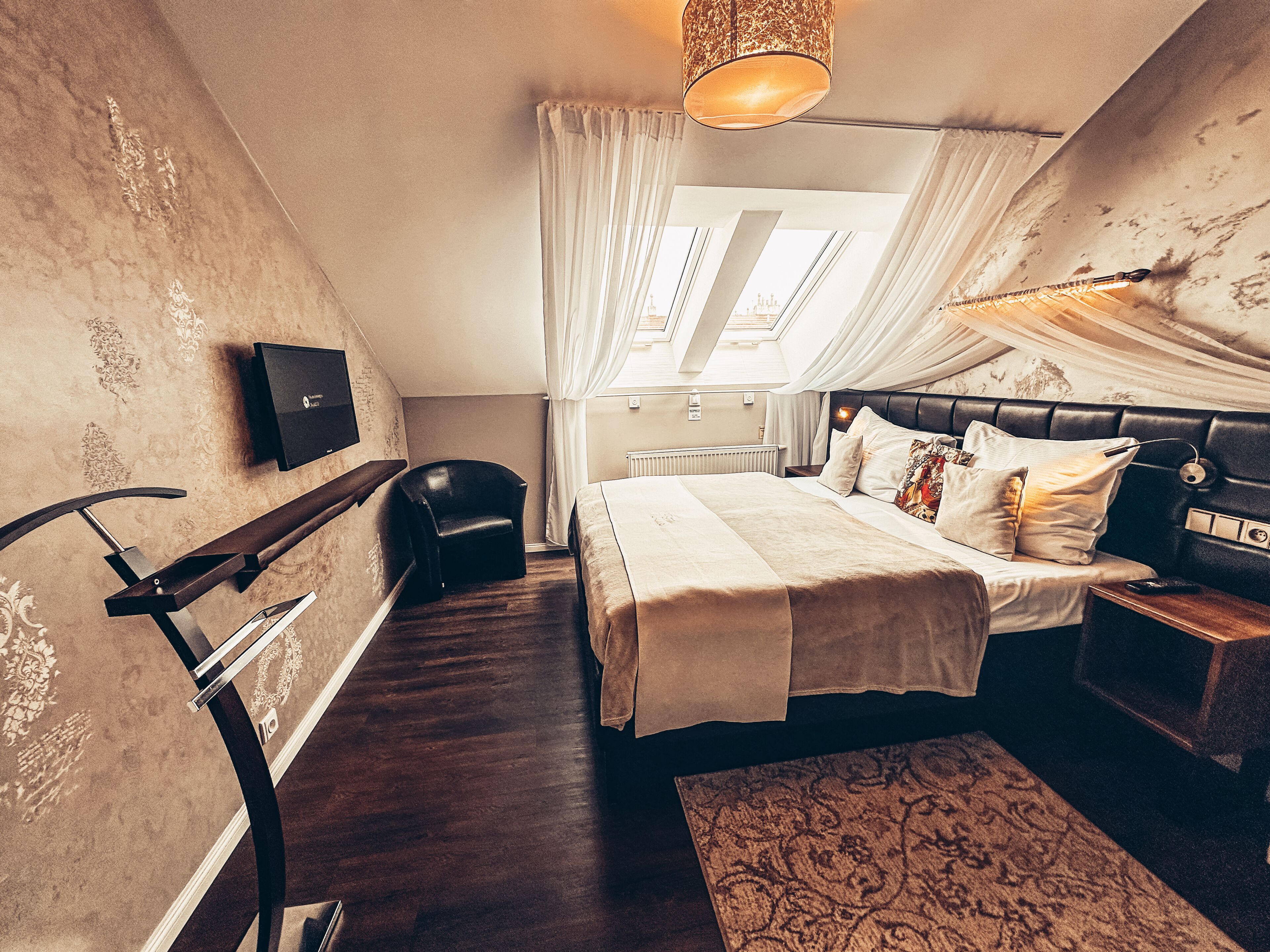 Un dormitorio con una cama, una silla, un televisor, un ventanal con cortinas y una alfombra estampada.