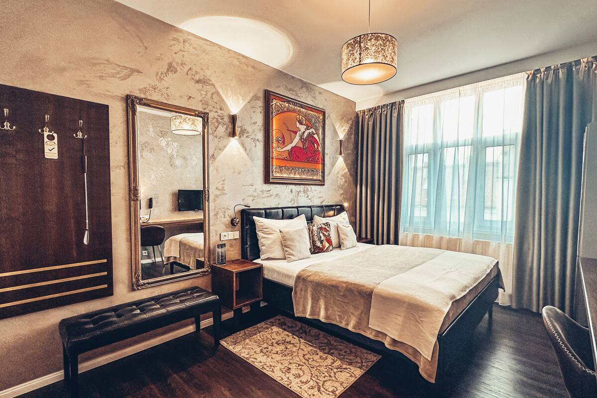 A bedroom with a large bed, a mirror, a bench, a rug, and a painting on the wall.