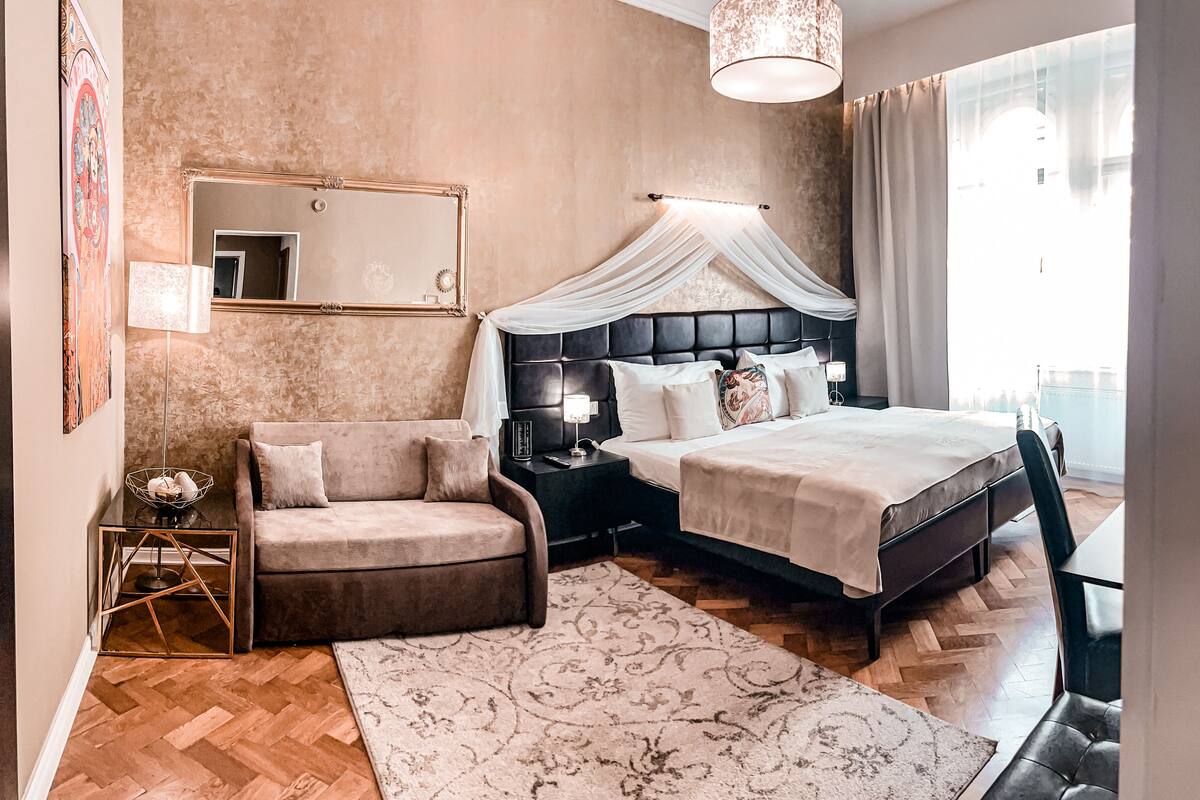 A modern bedroom with a large bed, a chaise lounge, a nightstand, and a mirror.
