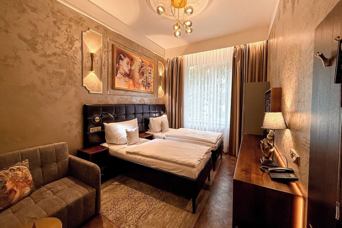 A hotel room with a double bed, a sofa, a desk with a telephone, and a large window with curtains.