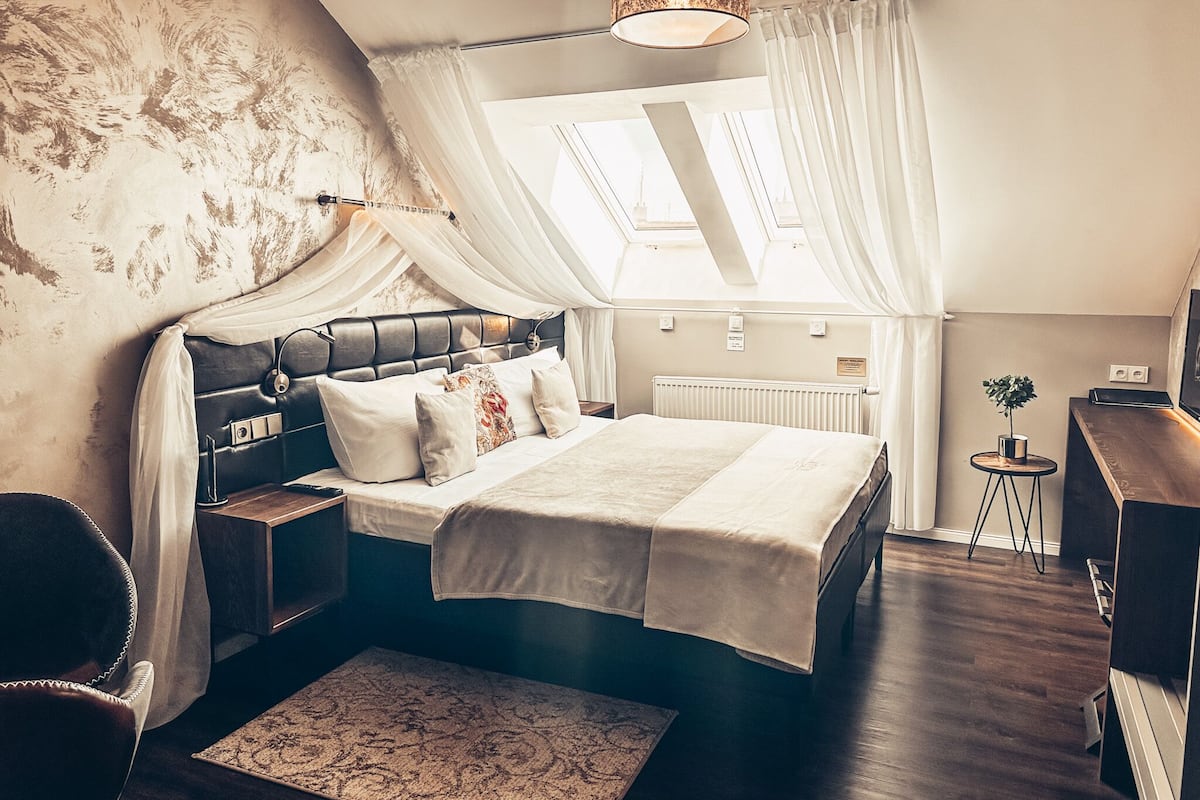 A bedroom with a large bed, a nightstand, a bedside table, a small round table with a plant, and a headboard with curtains.