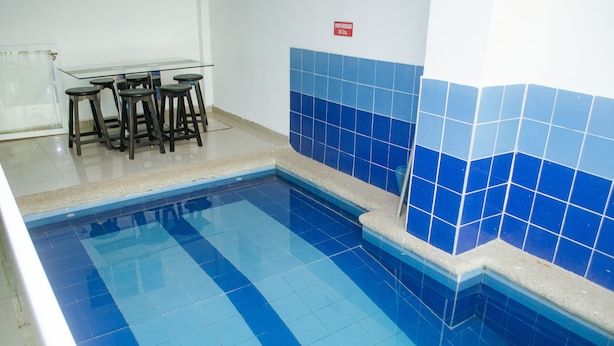 A swimming pool with blue tiles, a glass table with stools, and a tiled wall. - Hotel Piscina Real (Santa Marta)