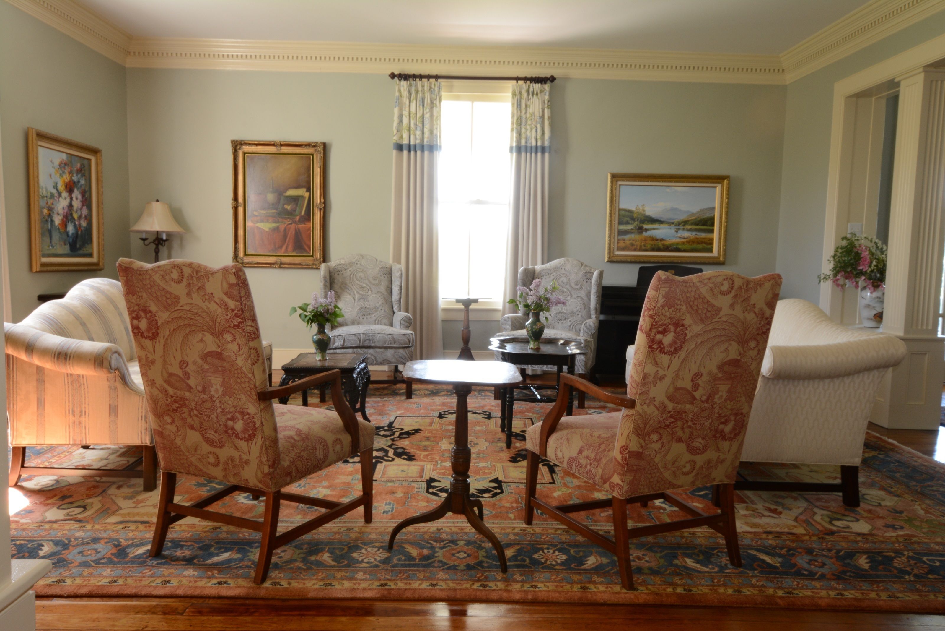 A traditional living room with patterned armchairs, a round coffee table, and a large window with curtains.