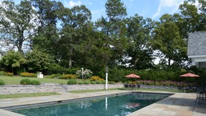 A rectangular pool with a stone border, surrounded by a patio area with outdoor furniture and umbrellas.