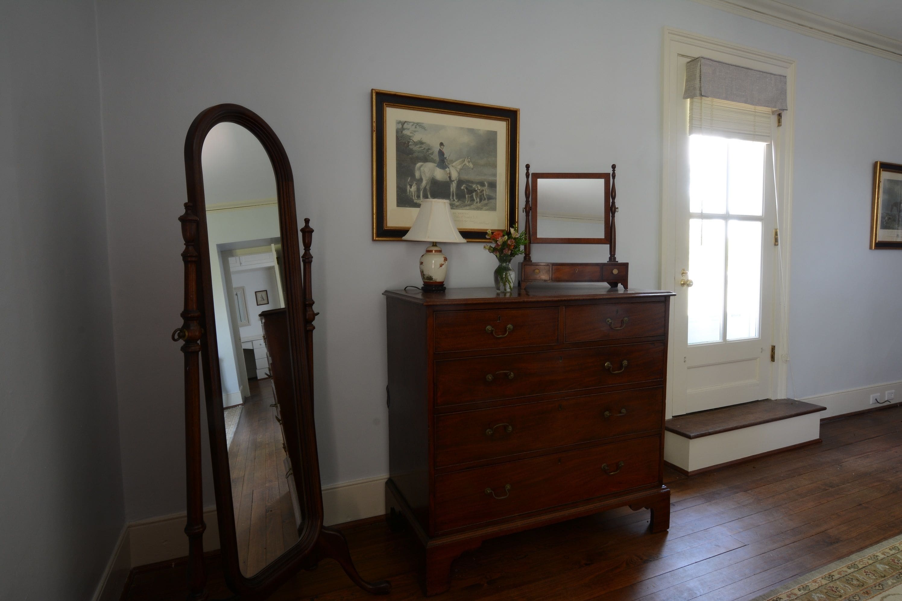 A room with a wooden dresser, a mirror, a framed picture on the wall, and a door with a window.