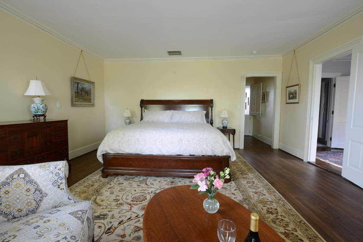 Een slaapkamer met een groot bed, een houten dressoir, een gestreept bankje, een klein tafeltje met een bloevas, en een fles champagne.