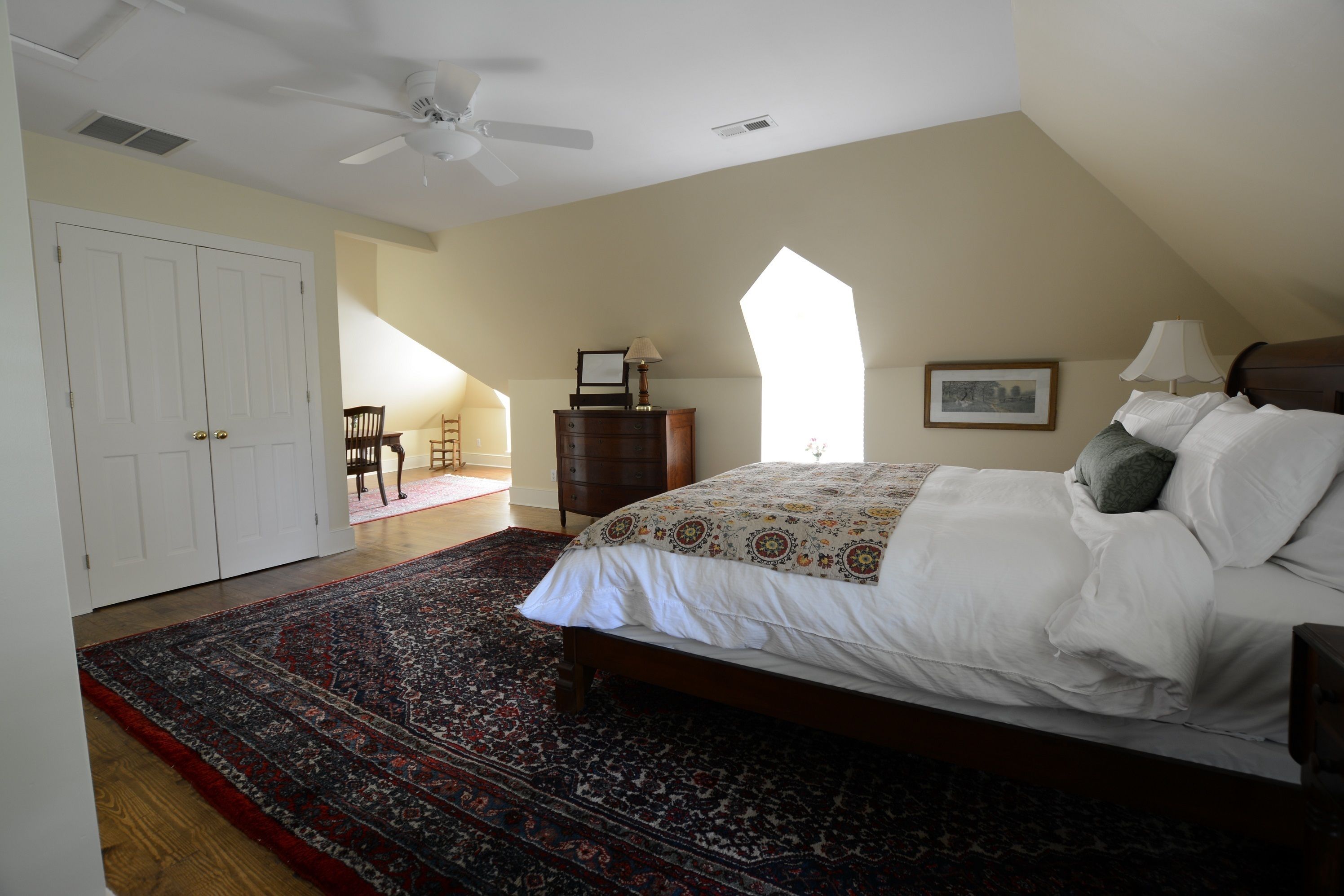 A bedroom with a large bed, a ceiling fan, a wooden dresser, a chair, and a large window.