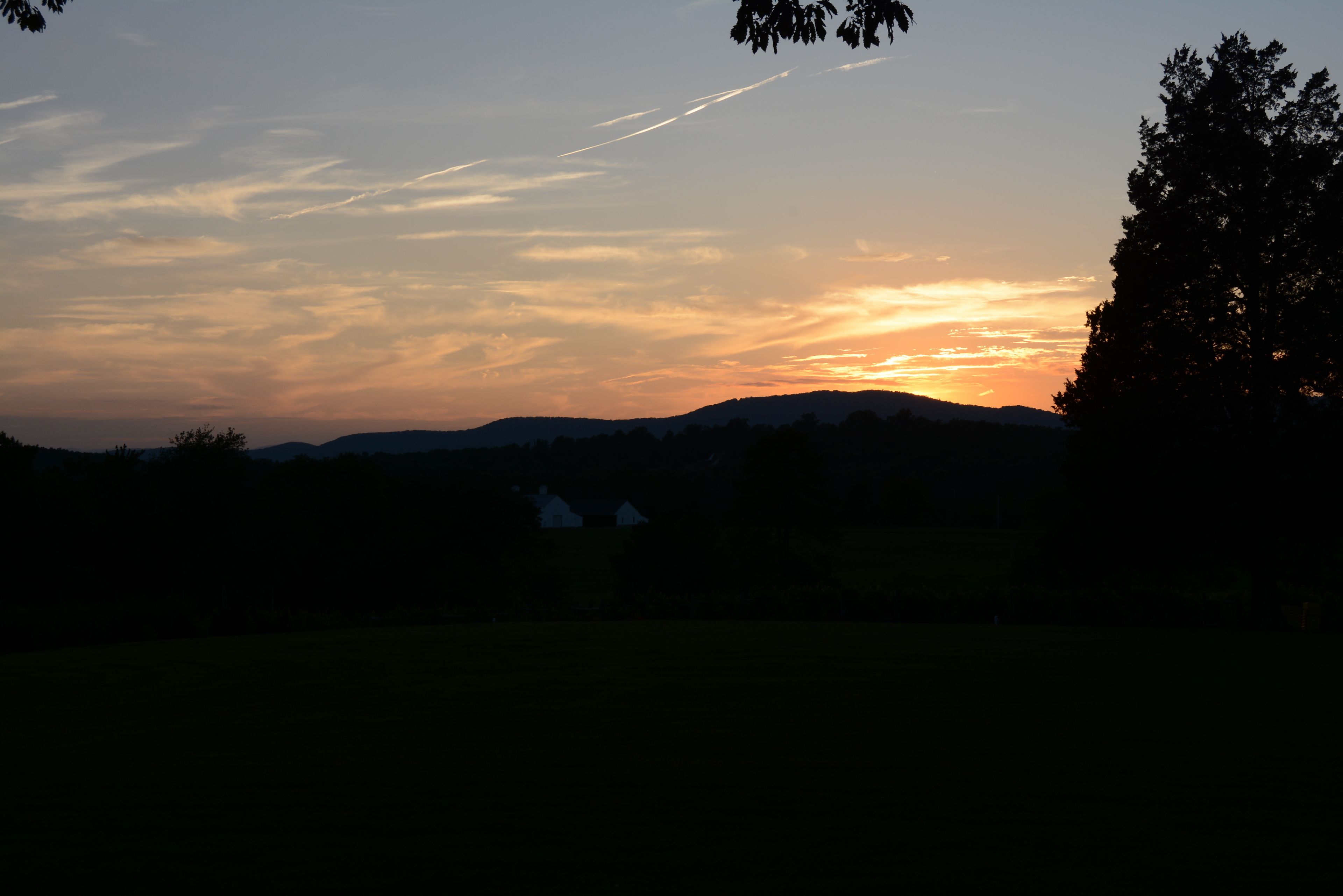 A sunset over a hilly landscape with silhouettes of trees and a house.