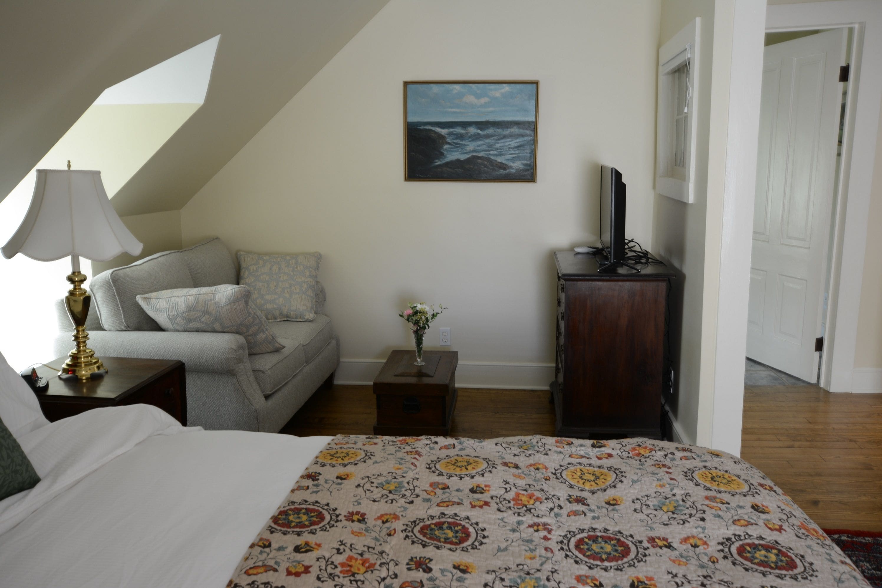 Une chambre à coucher avec un lit, un canapé, une petite table sur laquelle est posé un vase, un téléviseur sur un meuble et un tableau représentant la mer accroché au mur.