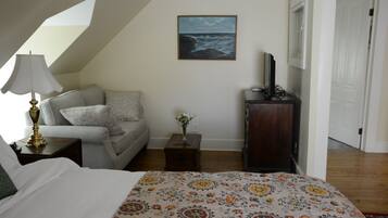 Une chambre à coucher avec un lit, un canapé, une petite table sur laquelle est posé un vase, un téléviseur sur un meuble et un tableau représentant la mer accroché au mur.