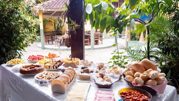 Una mesa de buffet con diferentes alimentos, como pan, pasteles y frutas, ubicada al aire libre con vegetación de fondo.