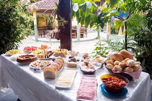 A buffet table with various food items, including bread, pastries, and fruits, set outdoors with greenery in the background. - Hospedagem La Bilbaina (Porto Seguro)