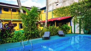 Una piscina con sillas de descanso, rodeada de abundante vegetación y un edificio con toldo rojo.