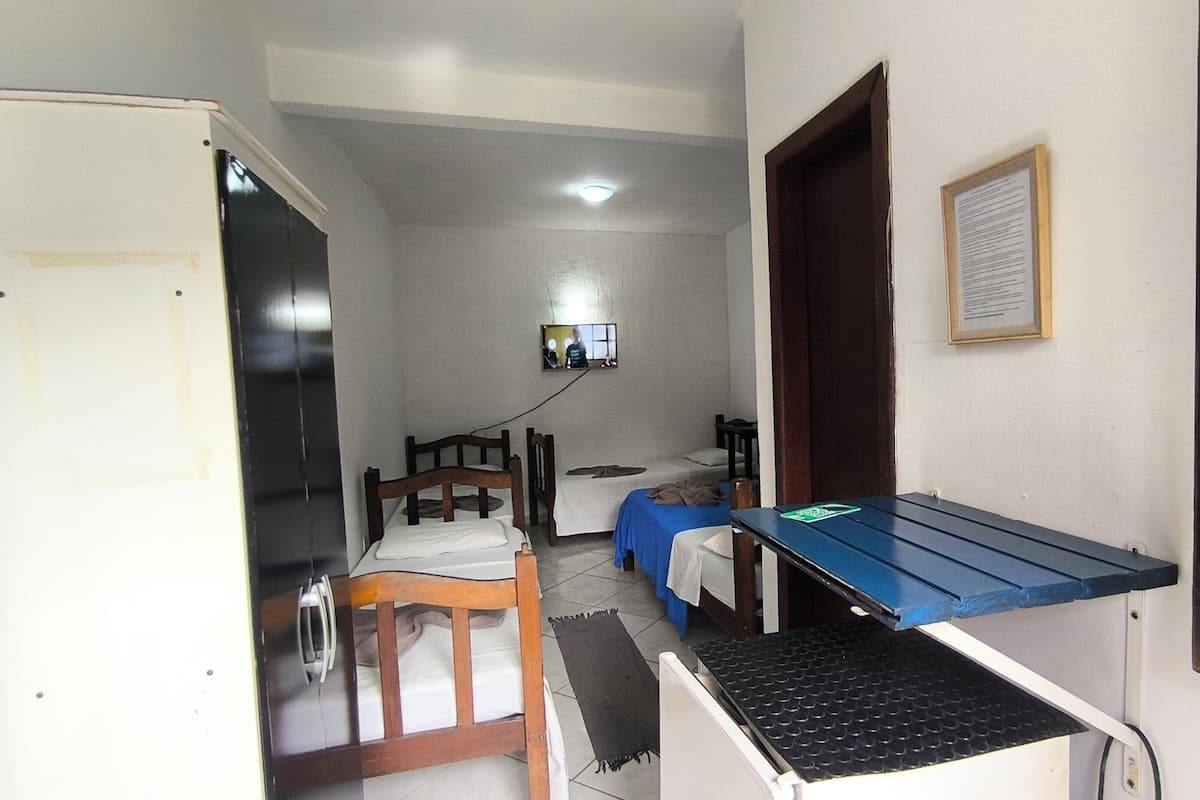 Quarto compacto com cama, escrivaninha, cadeira e televisão fixada na parede.