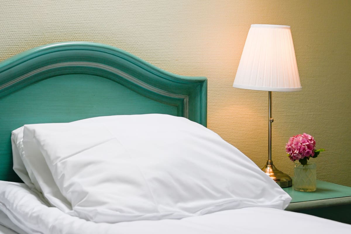Una cama bien hecha con sábanas blancas, un cabecero color turquesa y una lámpara de noche con una flor rosa.
