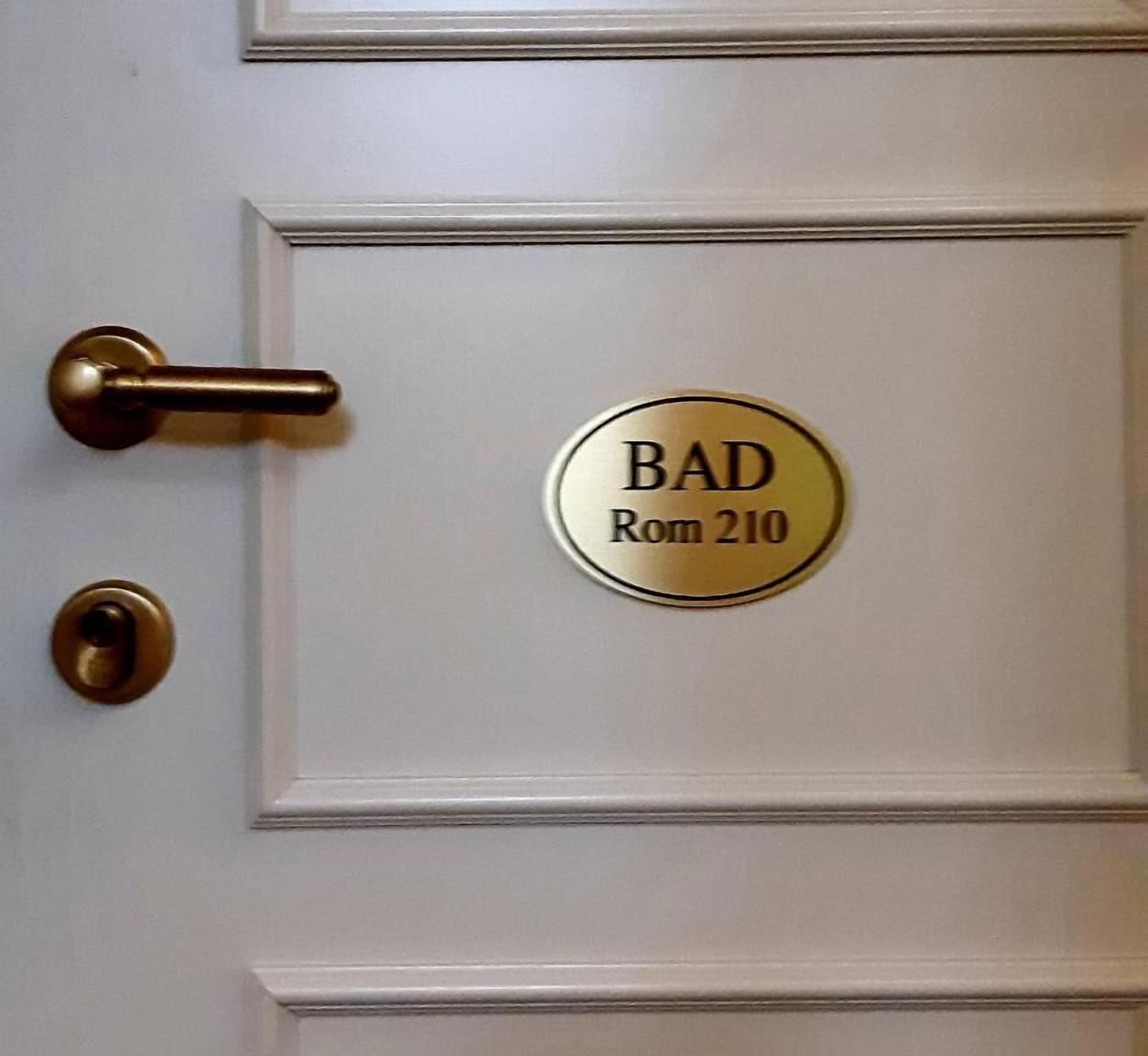 A door with a brass doorknob and a brass doorbell. There is a brass plaque with the inscription "BAD Rom 210".