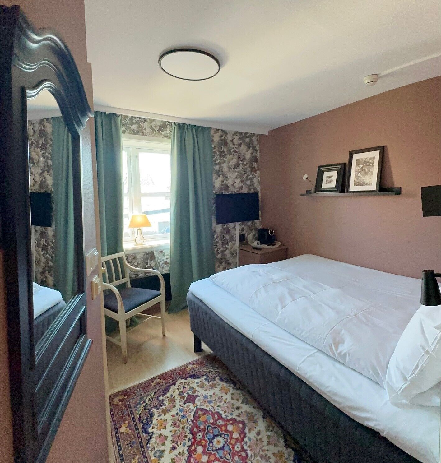A hotel room with a bed, a chair, a desk, a lamp, and a window with curtains.