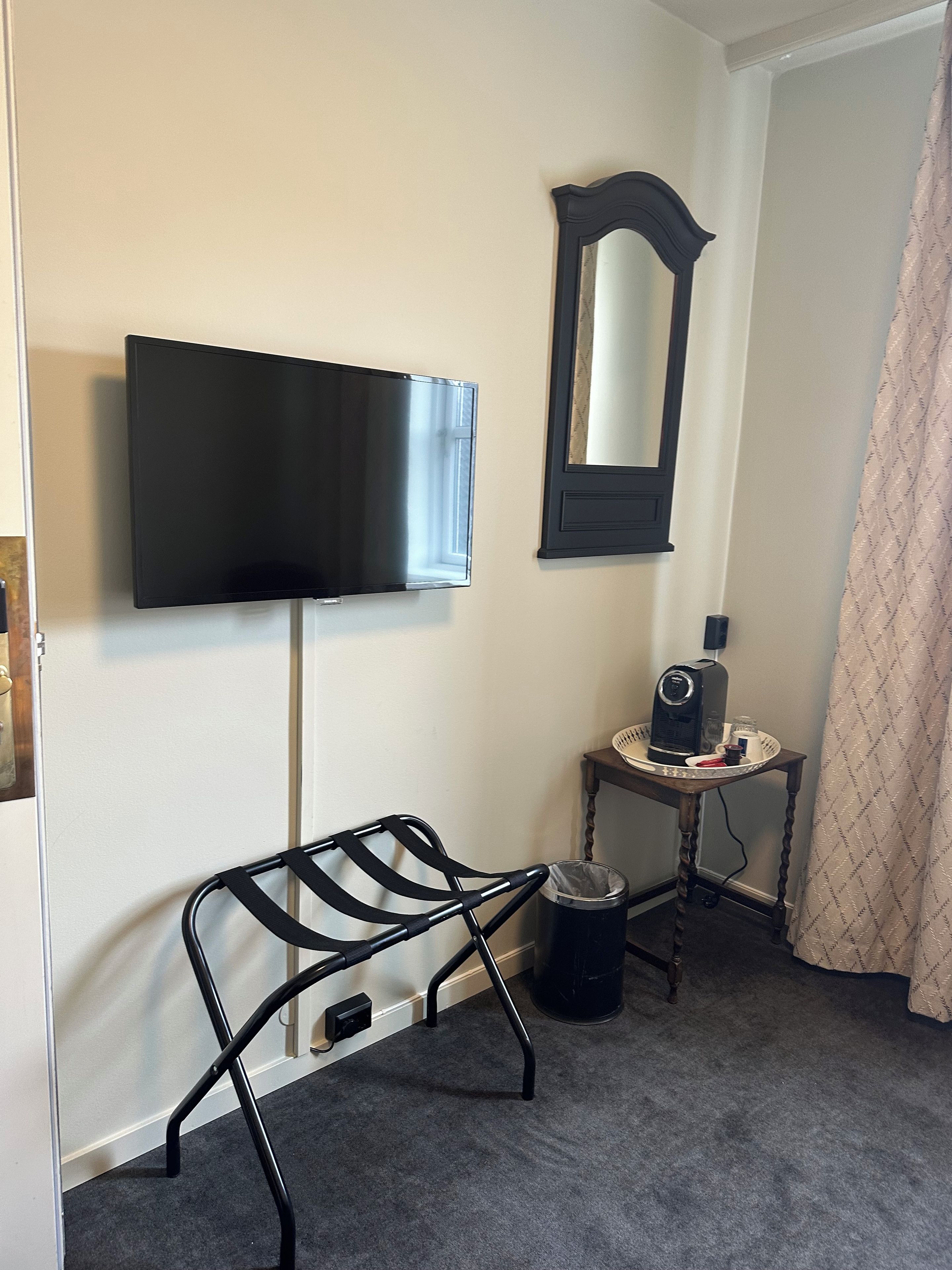 Een kamer met een wandtelevisie, een kleine tafel met een koffiezetapparaat, een prullenbak en een stoel met een metalen frame.