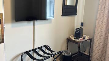 Een kamer met een wandtelevisie, een kleine tafel met een koffiezetapparaat, een prullenbak en een stoel met een metalen frame.