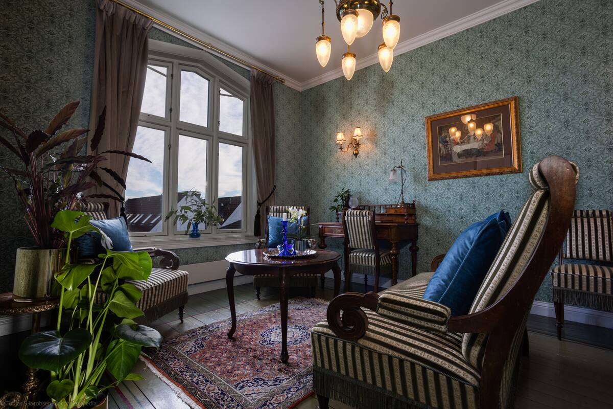Una habitación tradicional con papel tapiz estampado, un ventanal con cortinas, una araña y muebles clásicos, incluyendo un sillón a rayas y una mesa de madera.