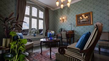 A traditional room with patterned wallpaper, a large window with curtains, a chandelier, and classic furniture including a striped armchair and a wooden table.