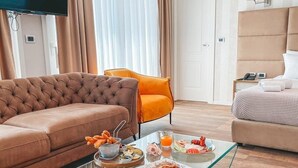 A modern hotel room with a brown sofa, orange armchair, and a glass coffee table with breakfast items.