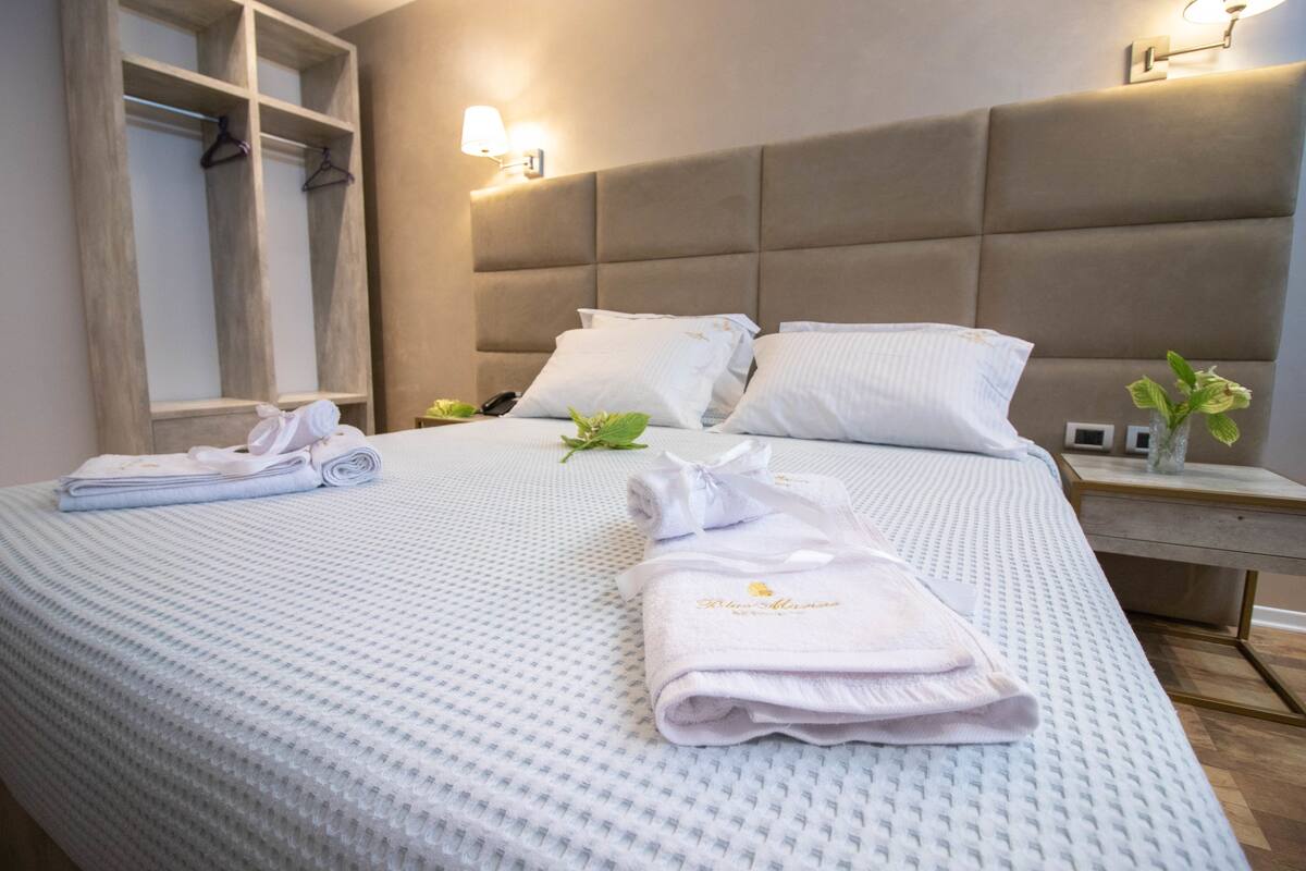 A neatly made bed with white linens and a folded towel, a bedside table with a lamp, and a wall-mounted light fixture.