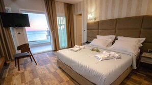 A hotel room with a large bed, a TV mounted on the wall, a chair, and a view of the sea through the window.