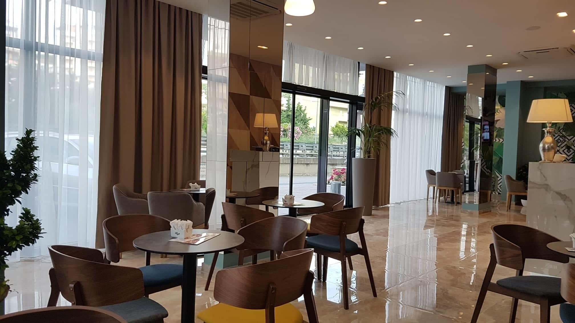 A modern hotel lobby with round tables and chairs, large windows with curtains, and a reception area with a TV.