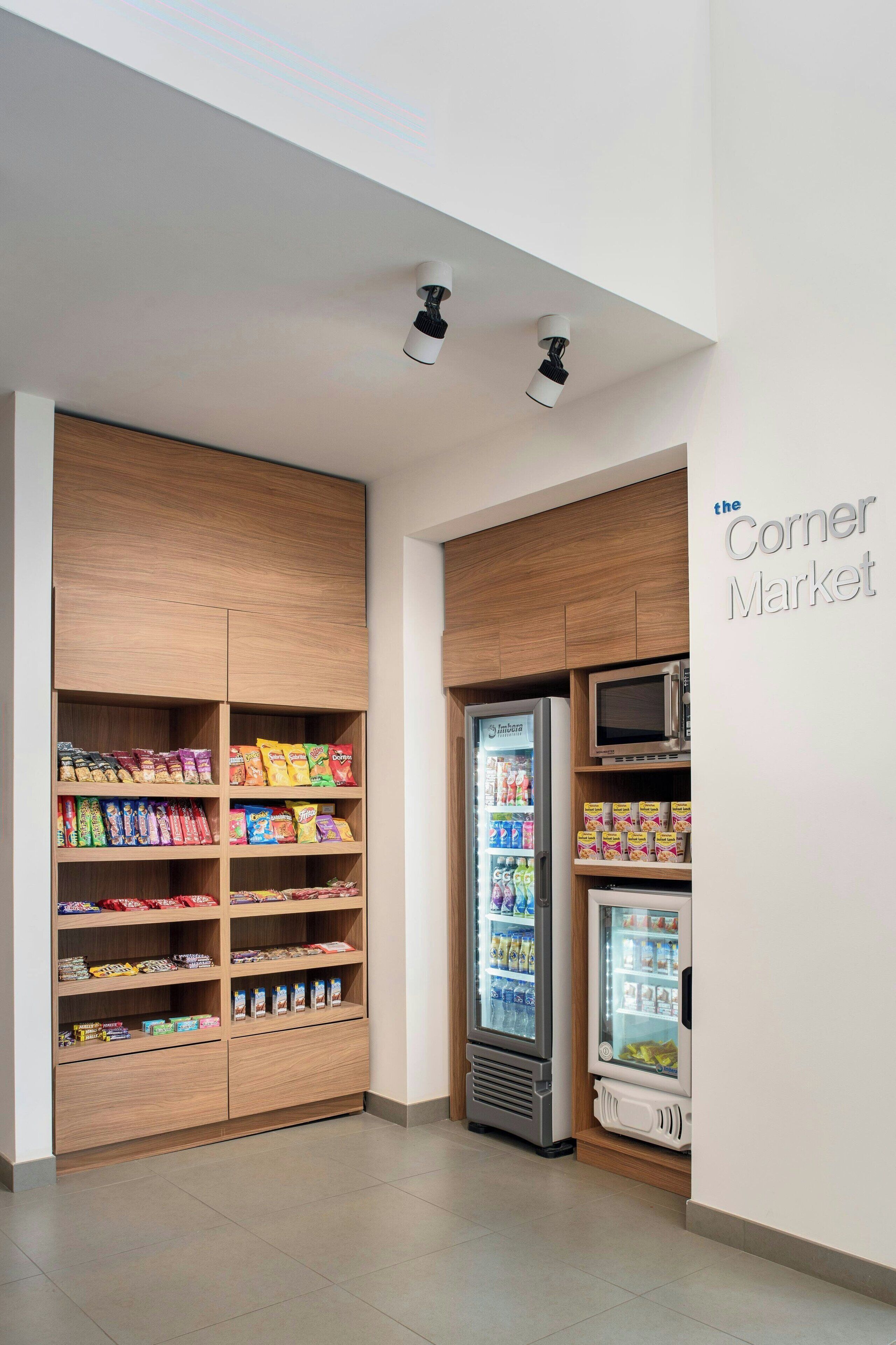 A corner market with wooden shelving stocked with snacks and beverages, a microwave, and a refrigerator.