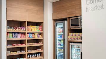 A corner market with wooden shelving stocked with snacks and beverages, a microwave, and a refrigerator.