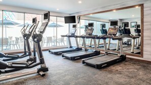 Uma academia com esteiras, elípticos e bicicletas de exercício, espelhos amplos e vista para área externa com mesas e cadeiras.