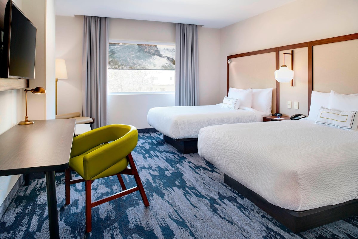 A hotel room with two beds, a desk, a chair, a TV, and a window with curtains.