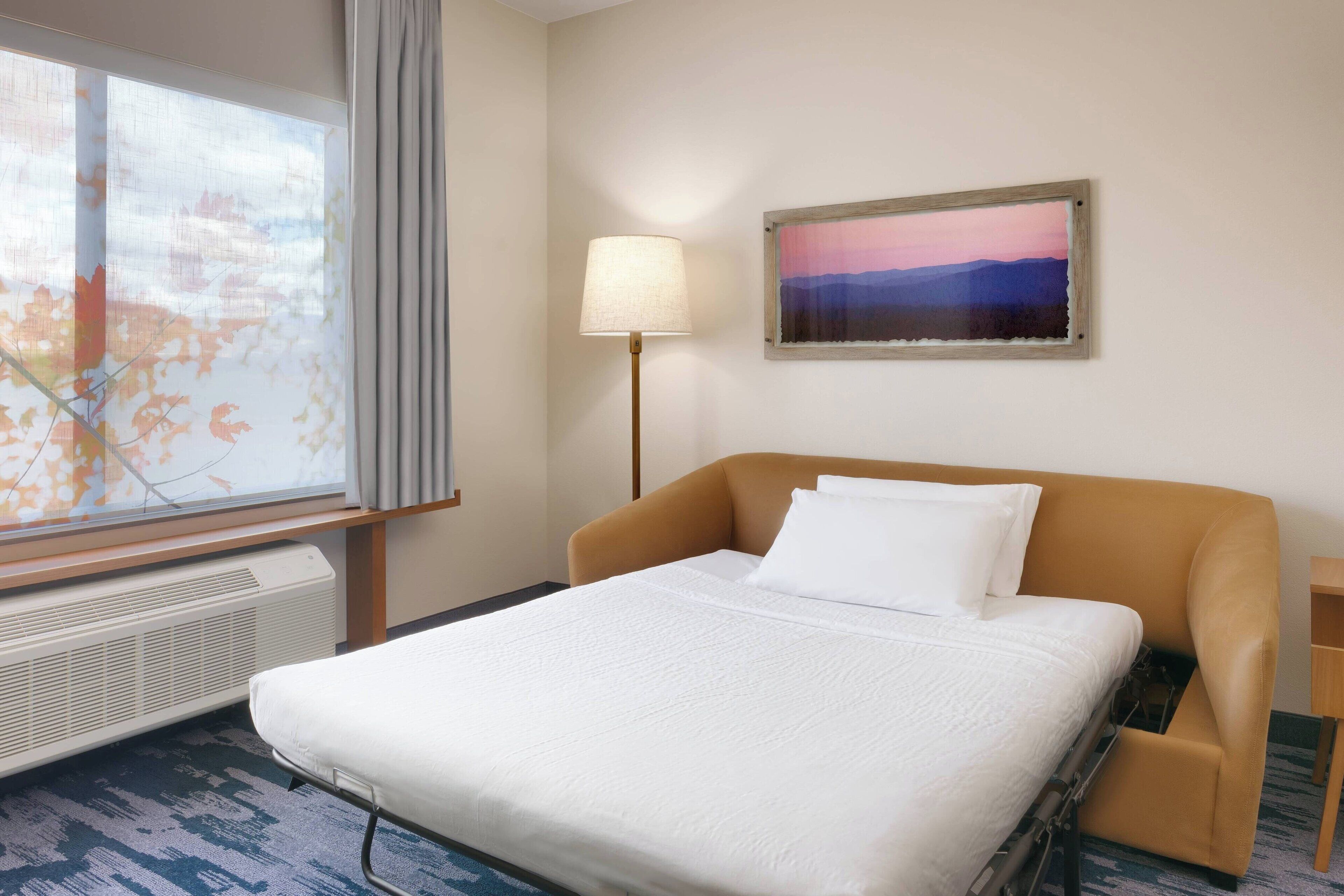 A hotel room with a bed, a window with a view of autumn leaves, a lamp, and a painting on the wall.