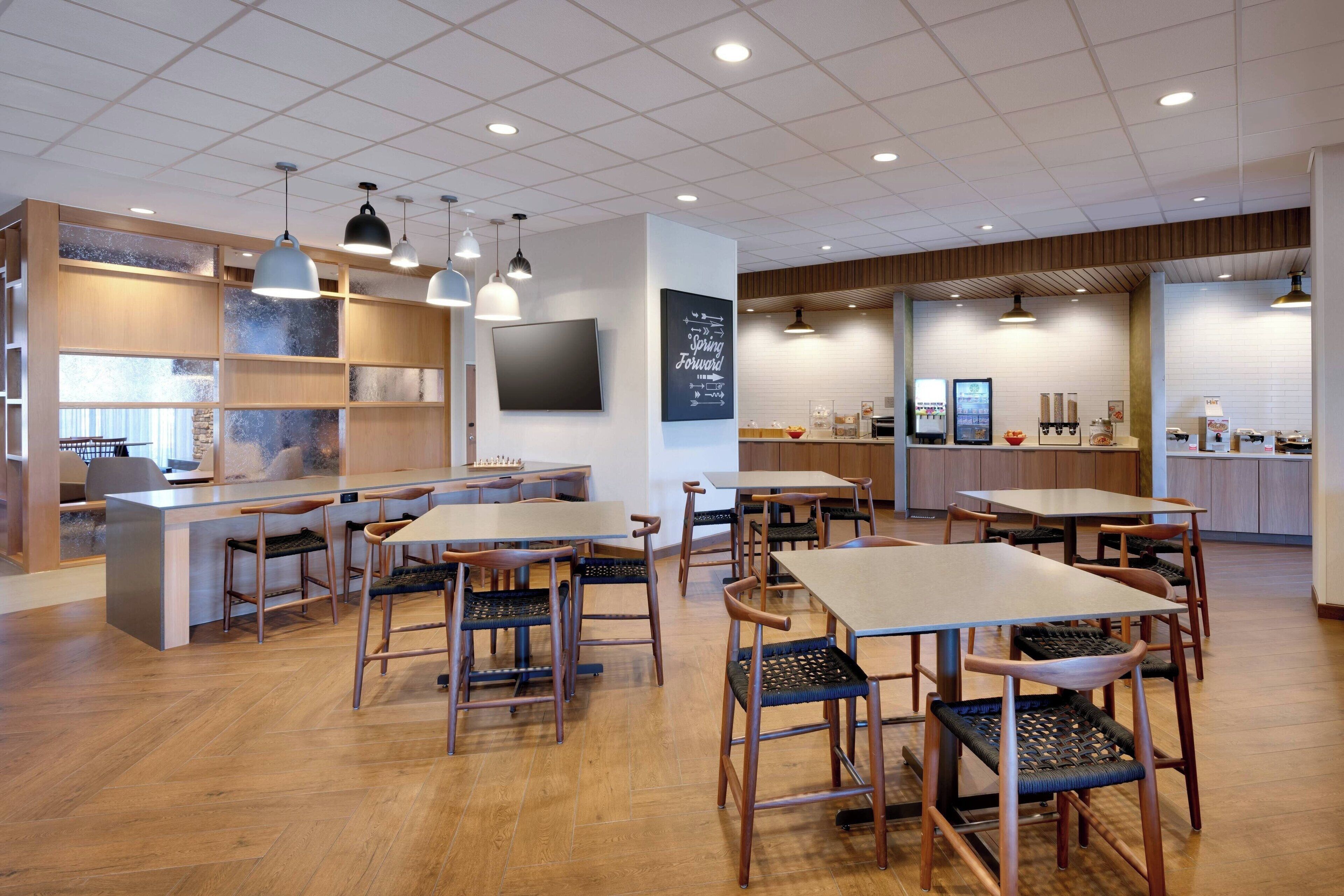 A modern restaurant with wooden flooring, tables, and chairs. There are hanging lights and a TV mounted on the wall.
