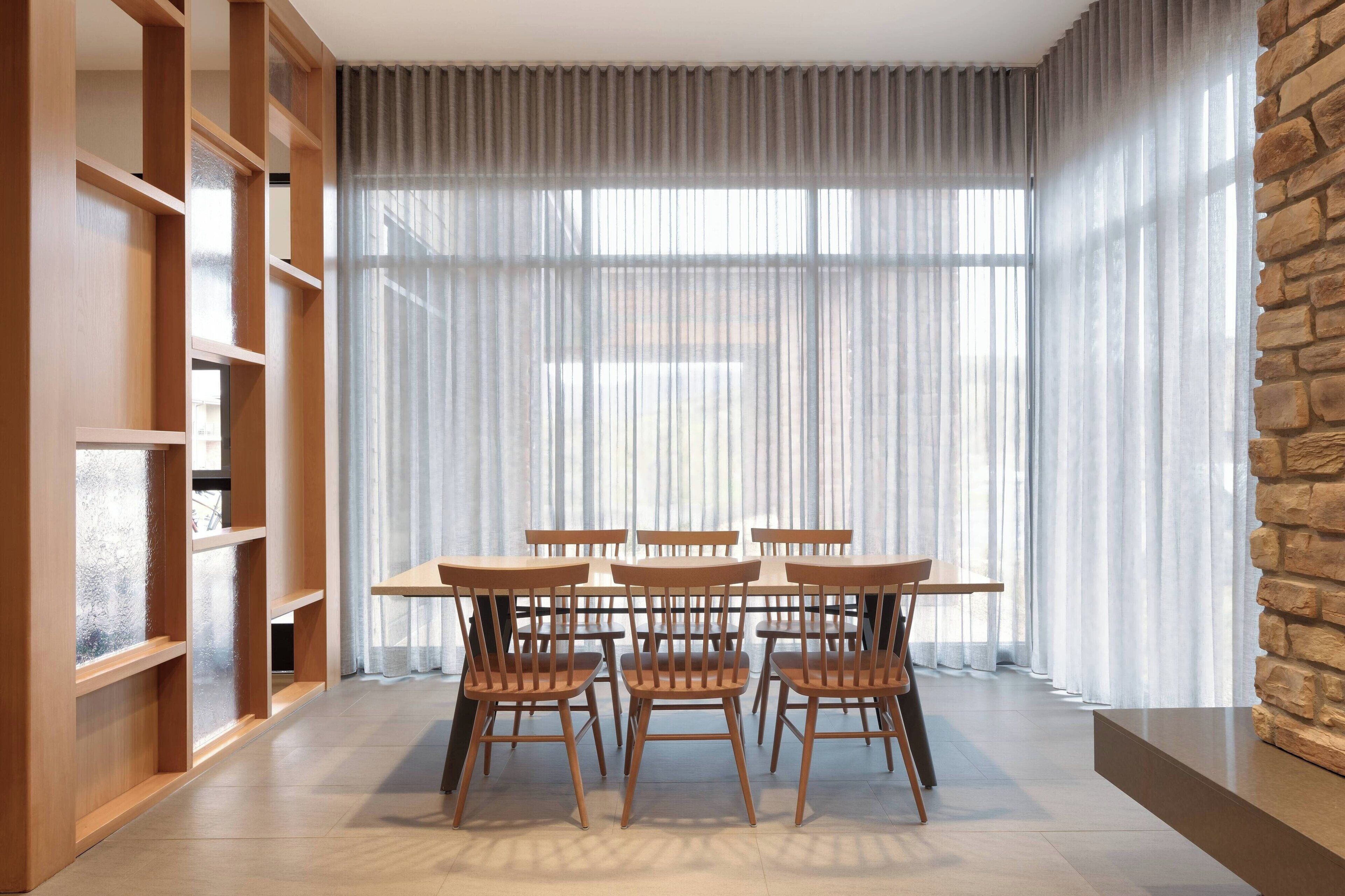 A dining area with a table and chairs, a wooden bookshelf, and a large window with sheer curtains.