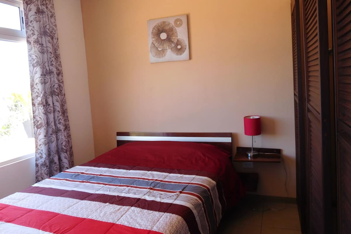 A bedroom with a bed, a nightstand, a lamp, a window with curtains, and a wall art piece.