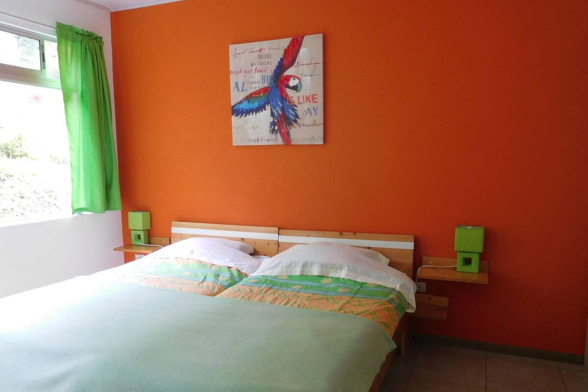 A bed with a green blanket, two pillows, and a colorful bird painting on the wall.