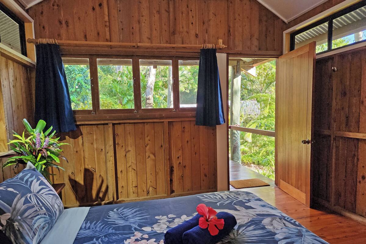 A wooden room with a bed, curtains, and a view of greenery outside.