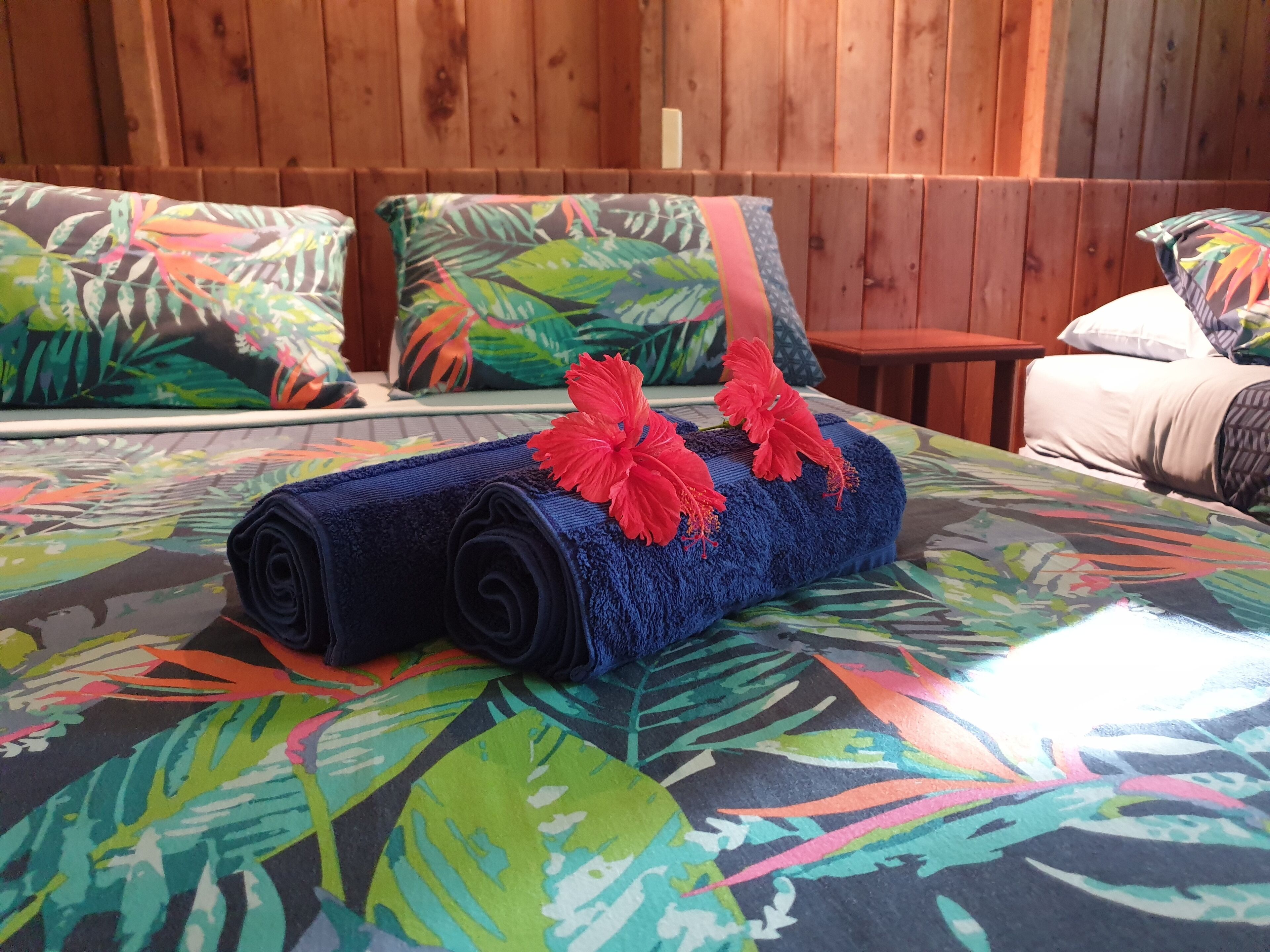 A bed with a rolled-up blue towel and red hibiscus flowers, set against a colorful leaf-patterned bedspread.